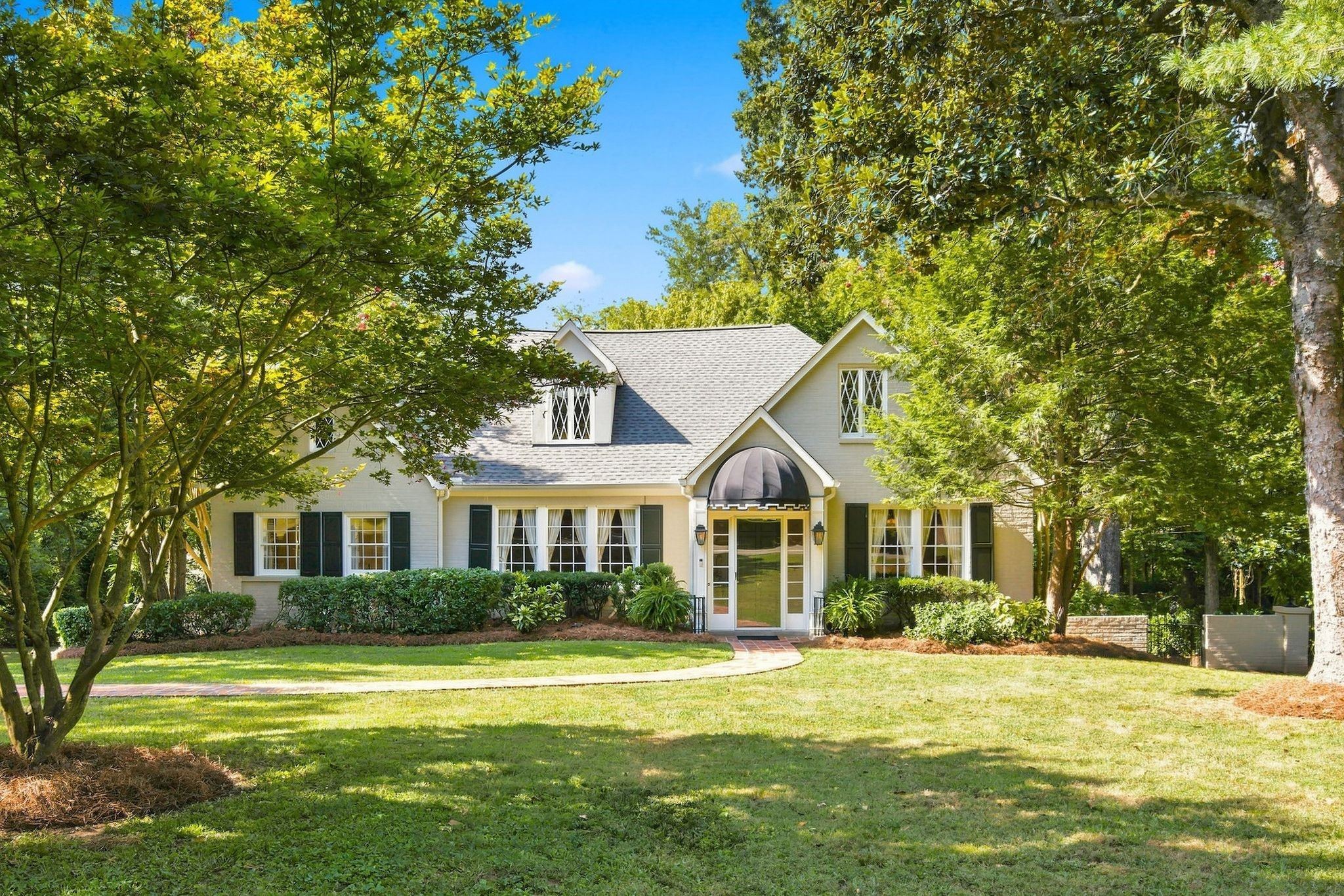 Front exterior of 317 Sutherland Avenue, a painted brick home with dormers, a brick walk and mature trees, one of the Belle Meade homes for sale in Nashville